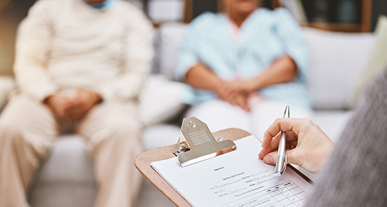 Closeup of a person holding a clipboard. A blurry couple on a couch is featured in the background 