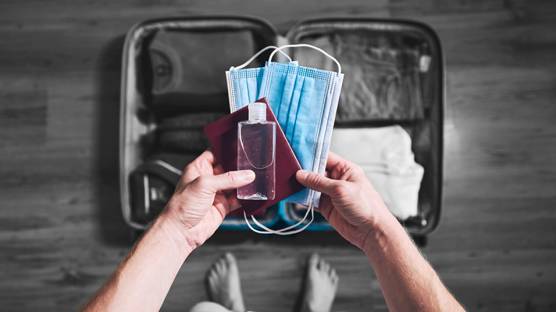 Open suitcase on floor and two hands holding hand sanitizer, a passport and medical masks. 