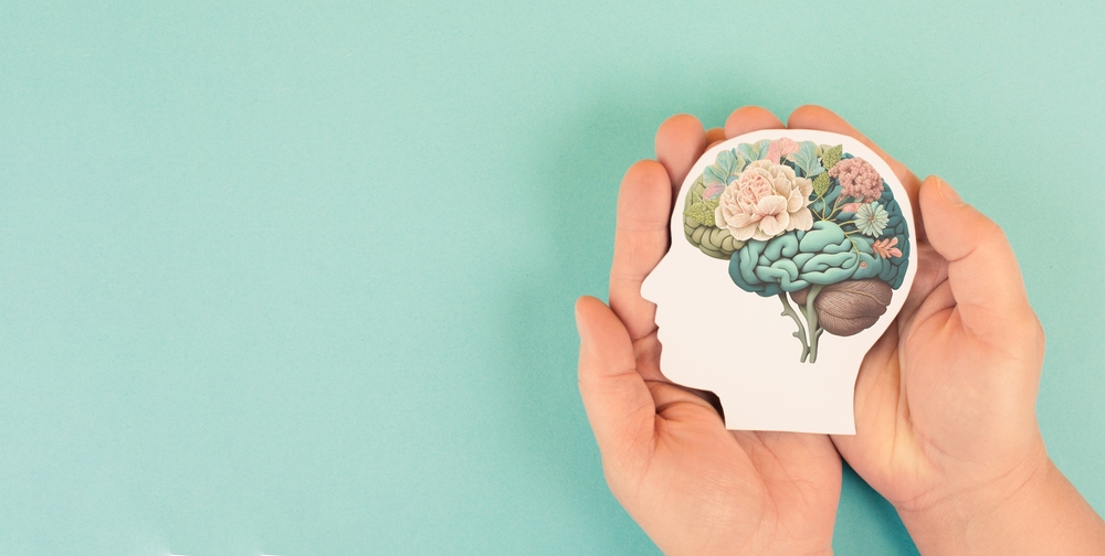 A pair of hands gently holding a paper cutout of a human head, with an artistic illustration of a brain filled with flowers, symbolizing mental health and well-being, against a pastel turquoise background. 